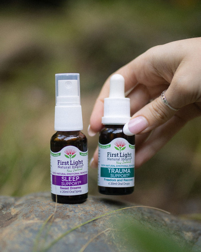 Two First Light essential oil bottles, Sleep and Trauma, held by a hand outdoors on a rock with a blurred forest background.